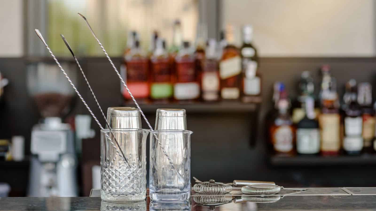 Two empty cocktail glasses with metal stirrers rest on a bar counter, hinting at the must-have mocktail mixers nearby. Blurred in the background are shelves stocked with various liquor bottles, alongside cocktail shakers and a strainer.