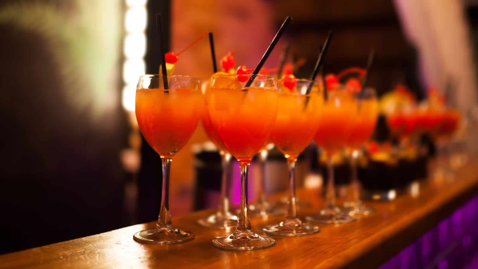 A row of cocktail glasses filled with a colorful orange and red drink, garnished with cherries and small straws, is placed on a wooden bar counter. Highlighting the must-have mocktail mixers, the background is softly blurred, emphasizing the bright drinks in the foreground.