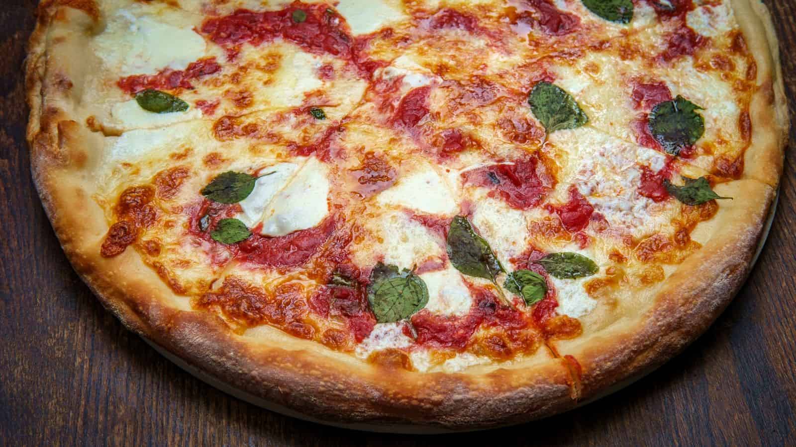 A close-up of a pizza with a golden-brown crust, topped with melted cheese, tomato sauce, and fresh basil leaves. The pizza is placed on a wooden surface.