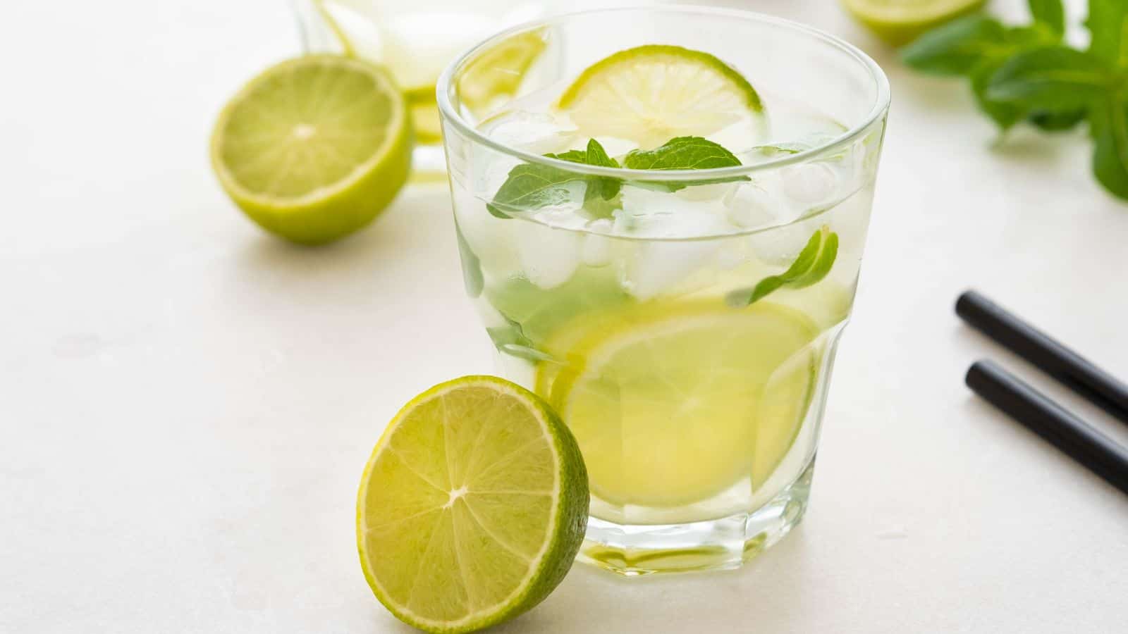 A glass of lime-infused beverage with ice and mint leaves. One slice of lime is in the drink, with another slice resting beside the glass. Additional lime and mint are in the background, alongside two black straws on a light surface.