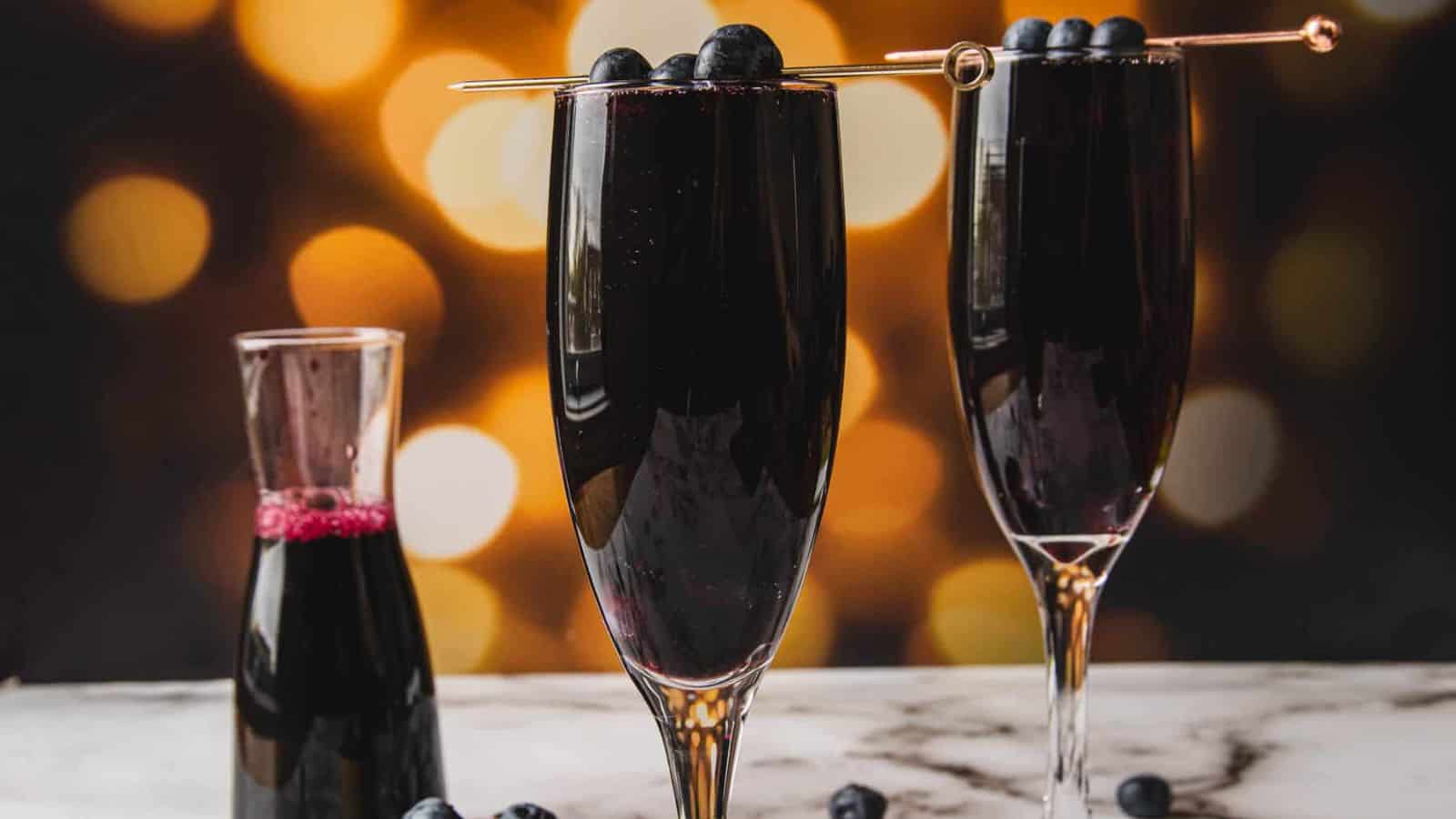 Two glasses of a dark red drink are on a marble surface, each garnished with a skewer of blueberries. A small bowl of blueberries and a glass pitcher with the same drink are in the background, with warm, blurred lights creating a cozy ambiance.