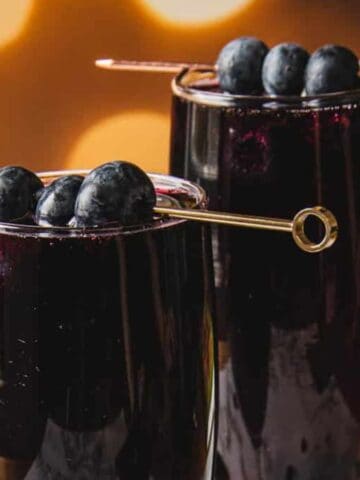 Two tall glasses filled with a dark purple beverage, each garnished with three blueberries on a skewer. The background is softly blurred with warm, glowing bokeh lights.