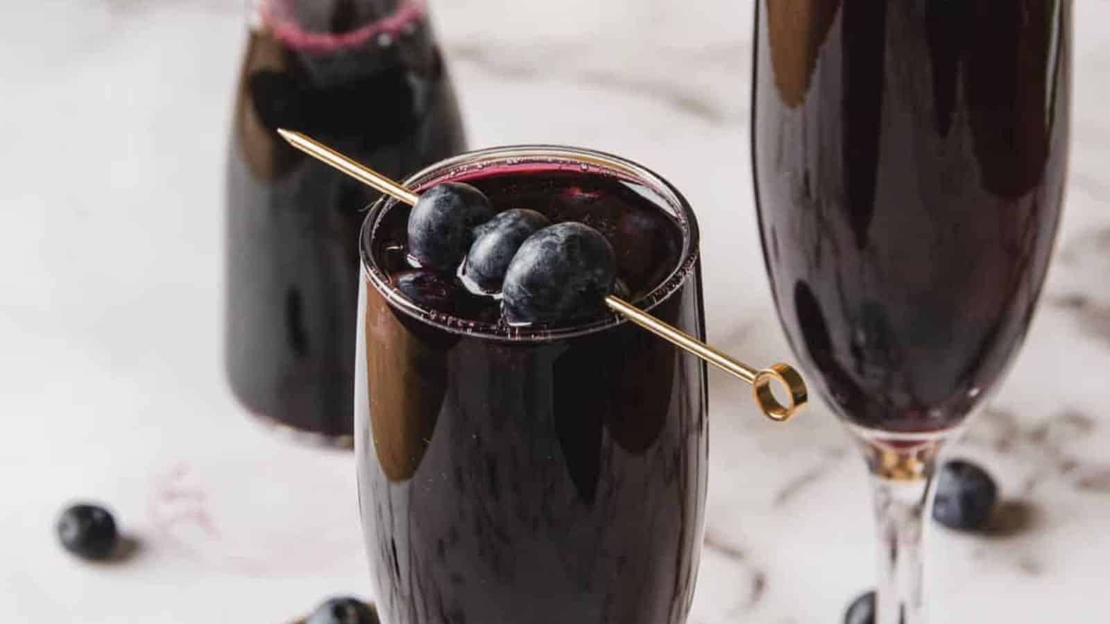 Two glasses filled with a dark purple drink are shown. One glass is garnished with blueberries on a skewer. More blueberries are scattered on the white surface, and an additional similar drink is partially visible in the background.