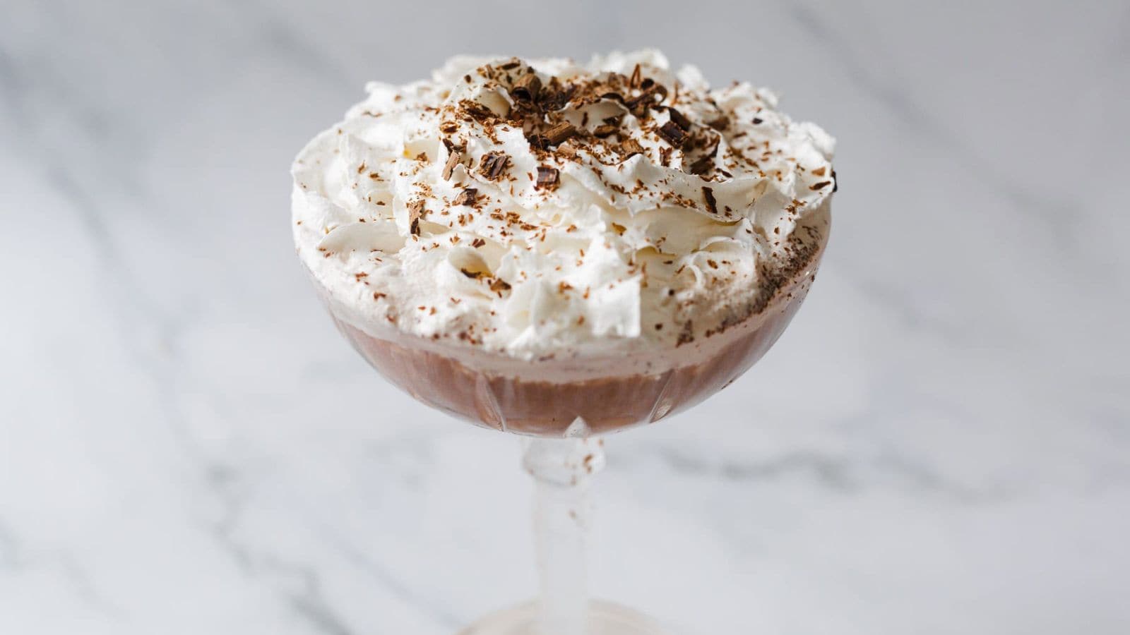 A chocolate dessert served in a glass bowl on a stand, topped with whipped cream and chocolate shavings, placed on a white marble surface.