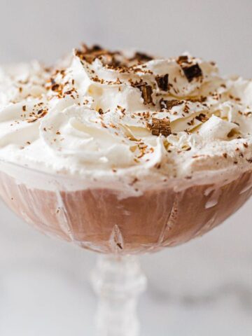 A creamy dessert in a glass dish, topped with fluffy whipped cream and sprinkled with chocolate shavings. The dish is set against a light, blurred background.