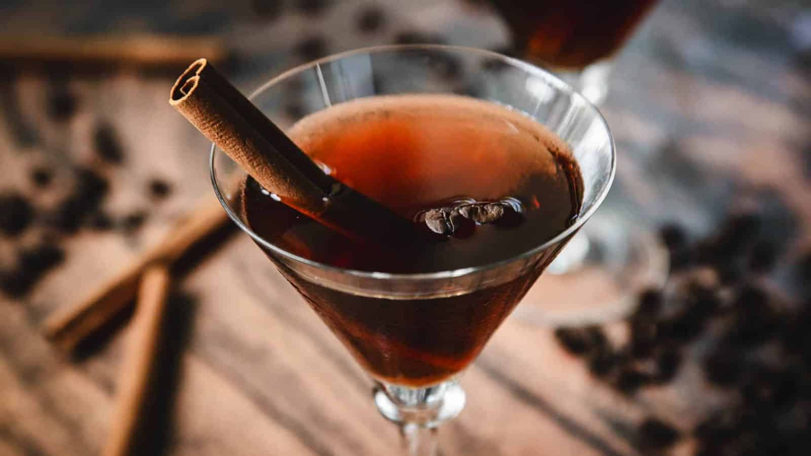 A close-up of a brown cocktail in a martini glass, garnished with a cinnamon stick. The background features scattered coffee beans and additional cinnamon sticks on a rustic wooden surface.