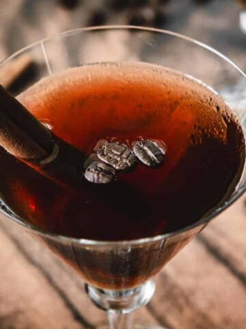 A martini glass filled with a dark brown cocktail, garnished with coffee beans and a cinnamon stick. Blurred cinnamon sticks and coffee beans are scattered on the wooden surface in the background.