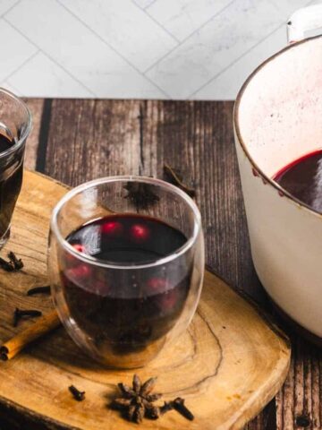 A white pot filled with mulled wine alongside two glass cups containing the beverage. The setup is on a wooden surface with spices like cinnamon sticks and star anise scattered nearby. A dark bottle is also present in the background.