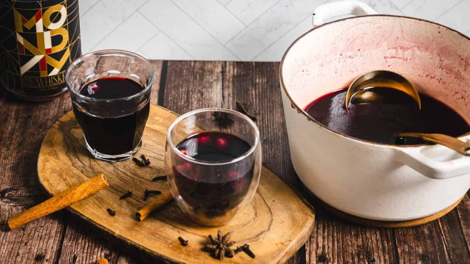 A glass of mulled wine sits on a wooden surface. The drink is garnished with a few floating berries. Nearby, there are whole cloves and a cinnamon stick on the wood. Another glass of dark liquid is partially visible in the background.