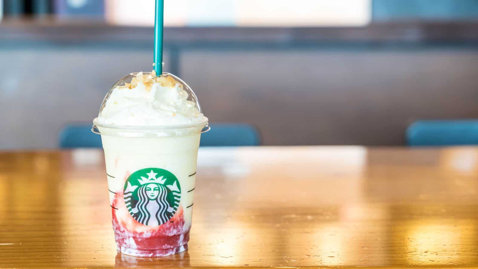 A Starbucks drink in a clear cup with a green straw sits on a wooden table. The beverage has a layer of red at the bottom and is topped with whipped cream under a plastic dome lid.