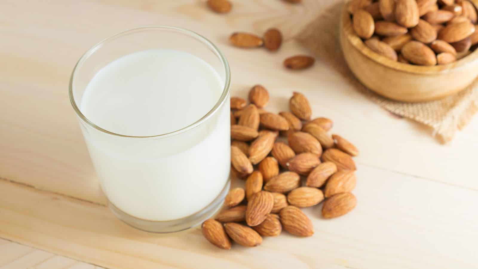 A glass of milk is placed on a wooden surface next to a cluster of almonds. A bowl filled with more almonds is visible in the background.