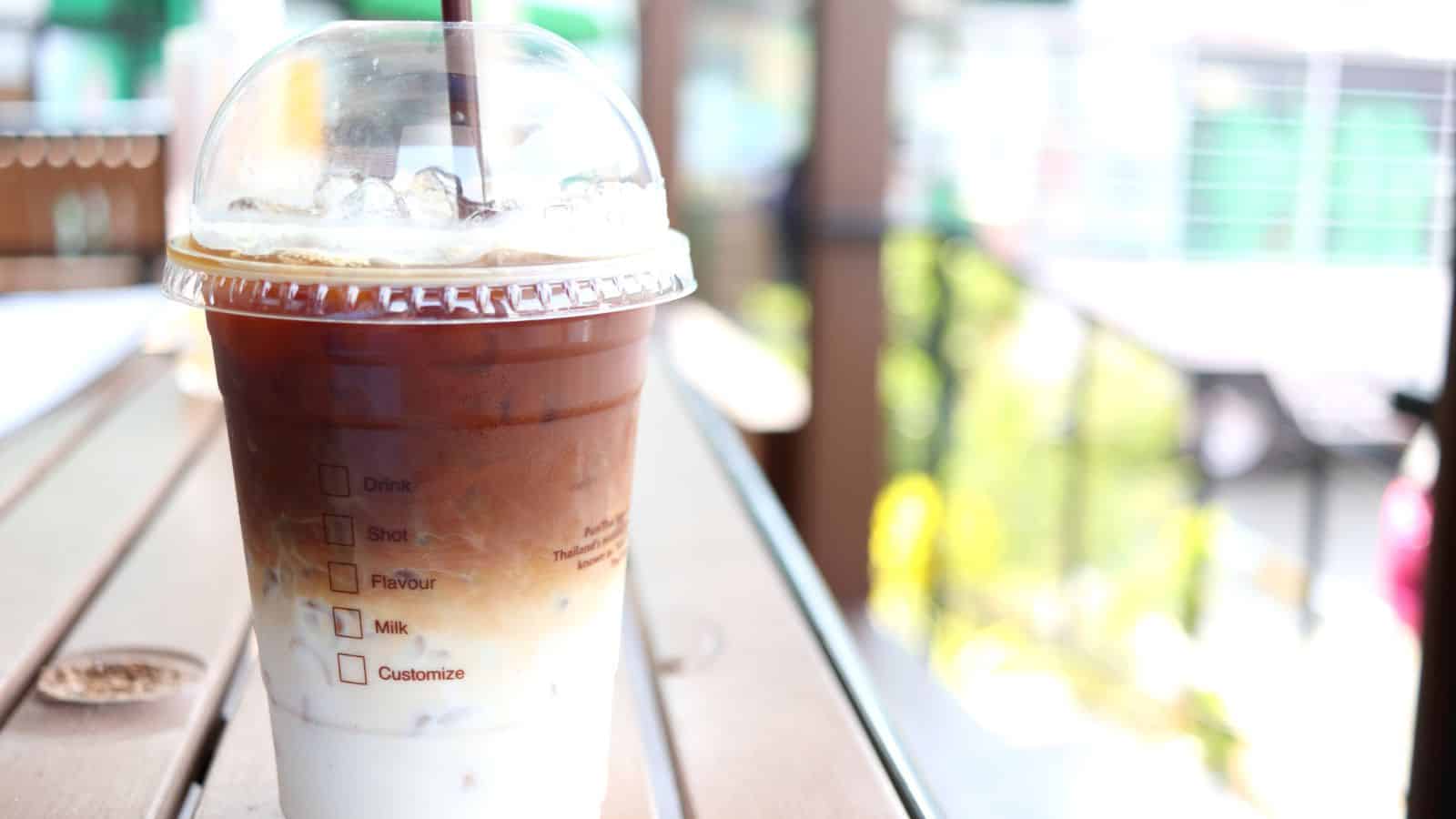 A clear plastic cup filled with iced coffee sits on a wooden table outdoors. The drink has visible layers of milk and coffee and is topped with ice. A brown straw protrudes from the domed lid. The background shows blurred greenery.