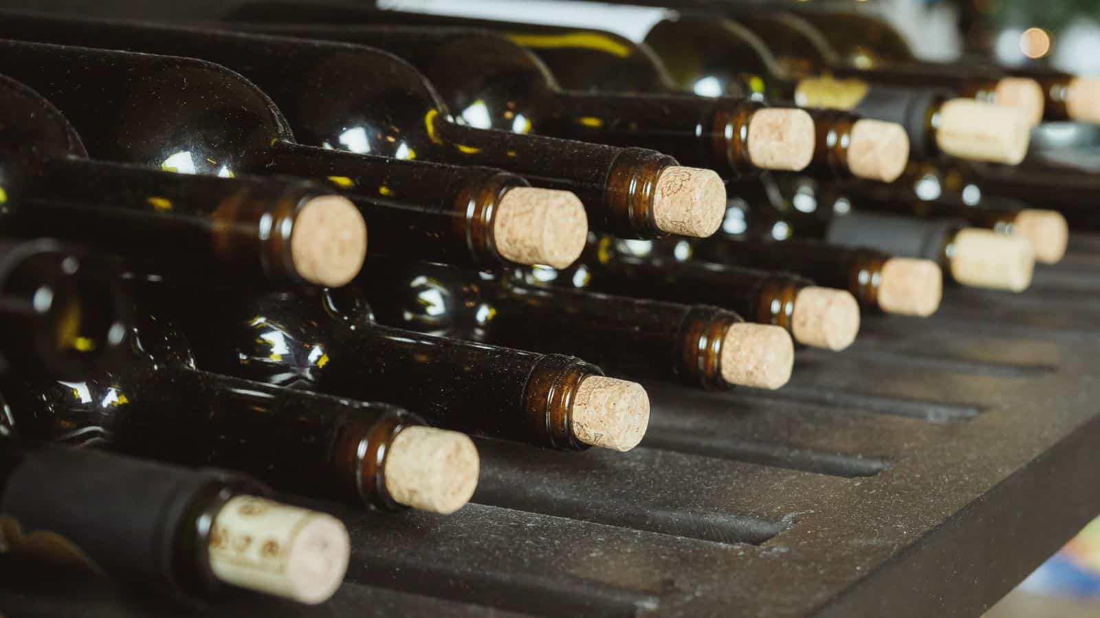 Rows of wine bottles with cork stoppers are placed horizontally in a rack. The bottles are dark green and arranged neatly, with the necks facing outward. The scene suggests a wine storage area.