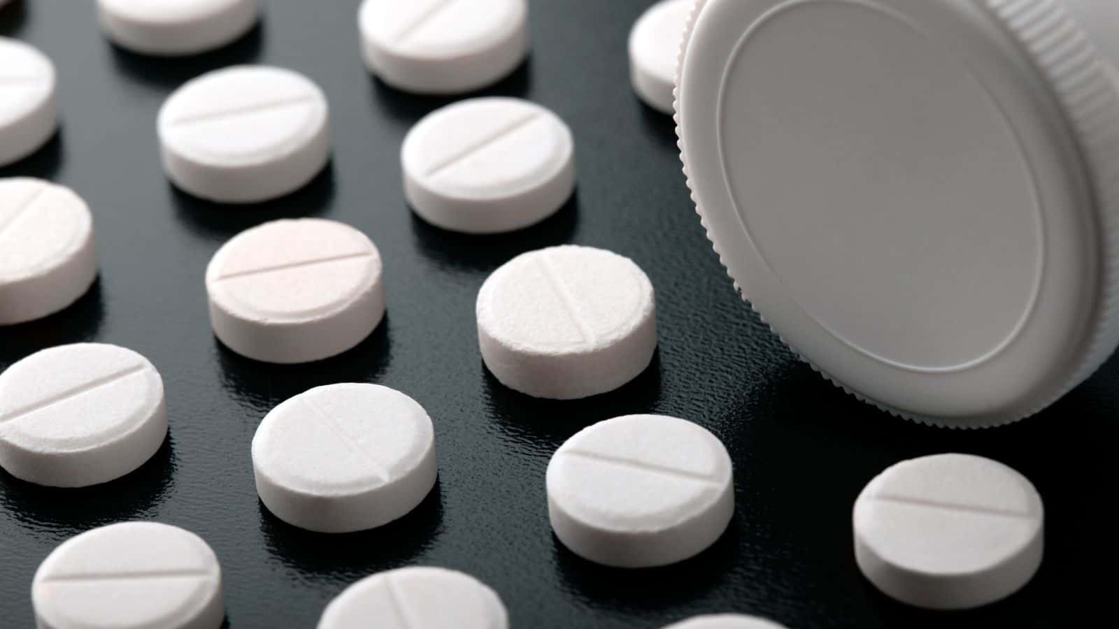 White round tablets with a scored line in the center are scattered on a dark surface next to an opened white bottle lying on its side. The arrangement is orderly, with the tablets placed in rows.