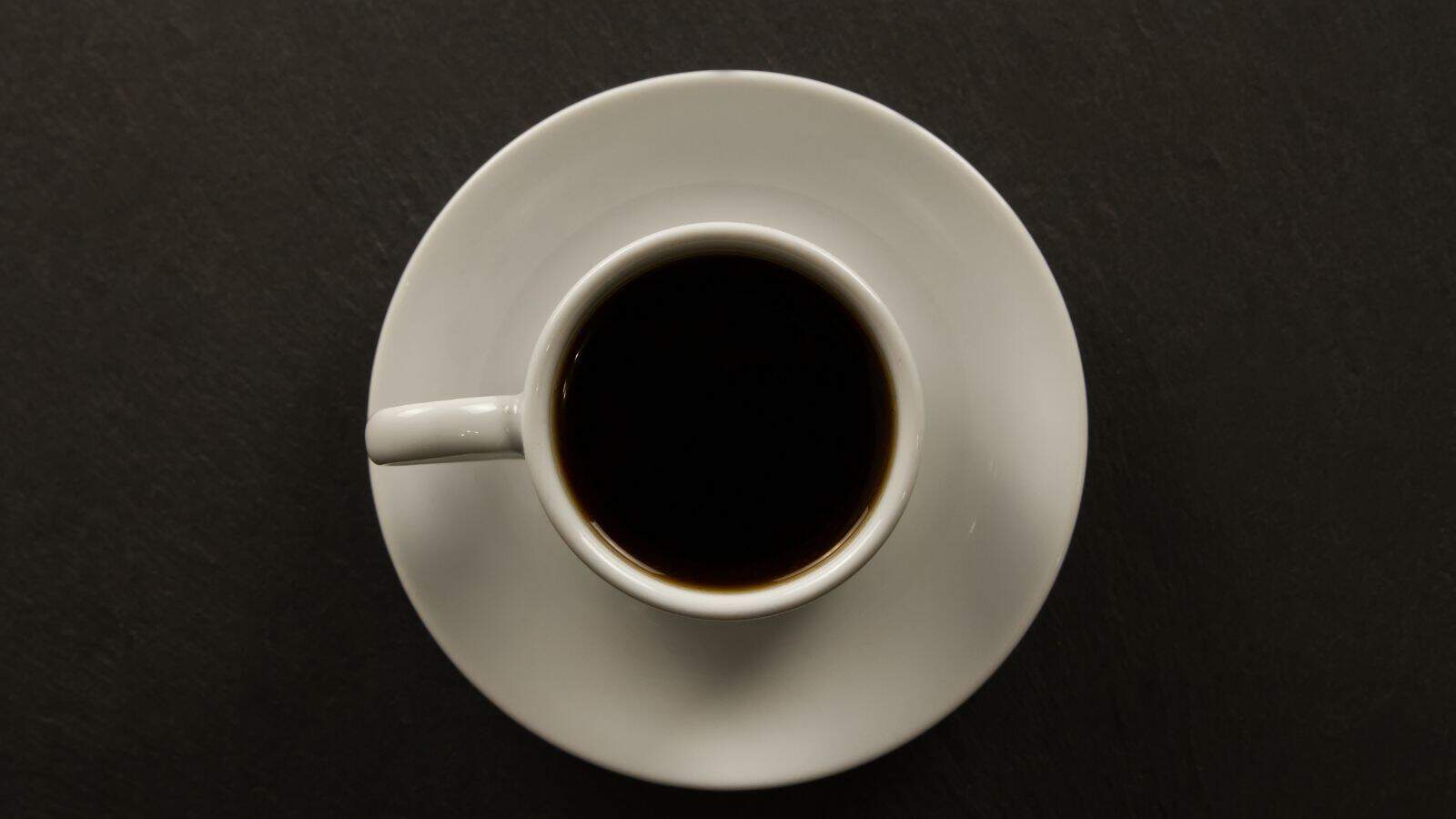 A white cup filled with black coffee sits on a matching white saucer against a dark surface, viewed from above.