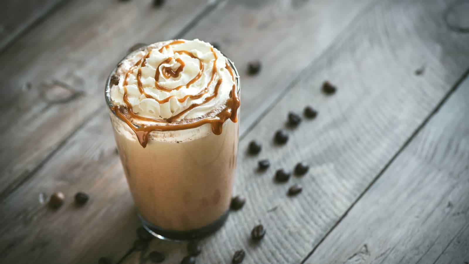 A glass of iced coffee topped with whipped cream and drizzled caramel sits on a wooden surface, surrounded by scattered coffee beans.