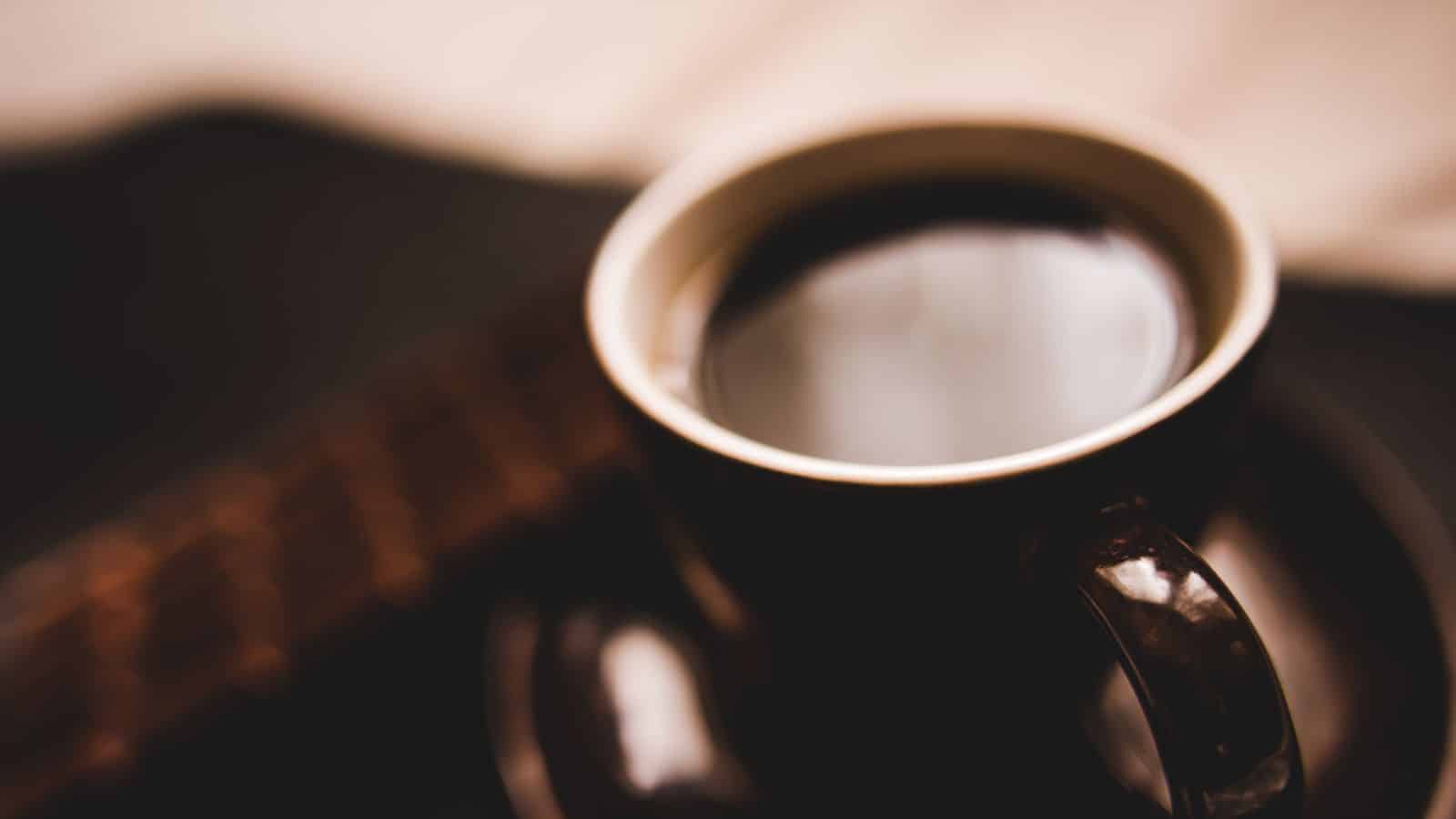 A close-up of a dark, steaming cup of coffee on a saucer, placed on a dark surface. The background is softly blurred, creating a warm and inviting atmosphere.