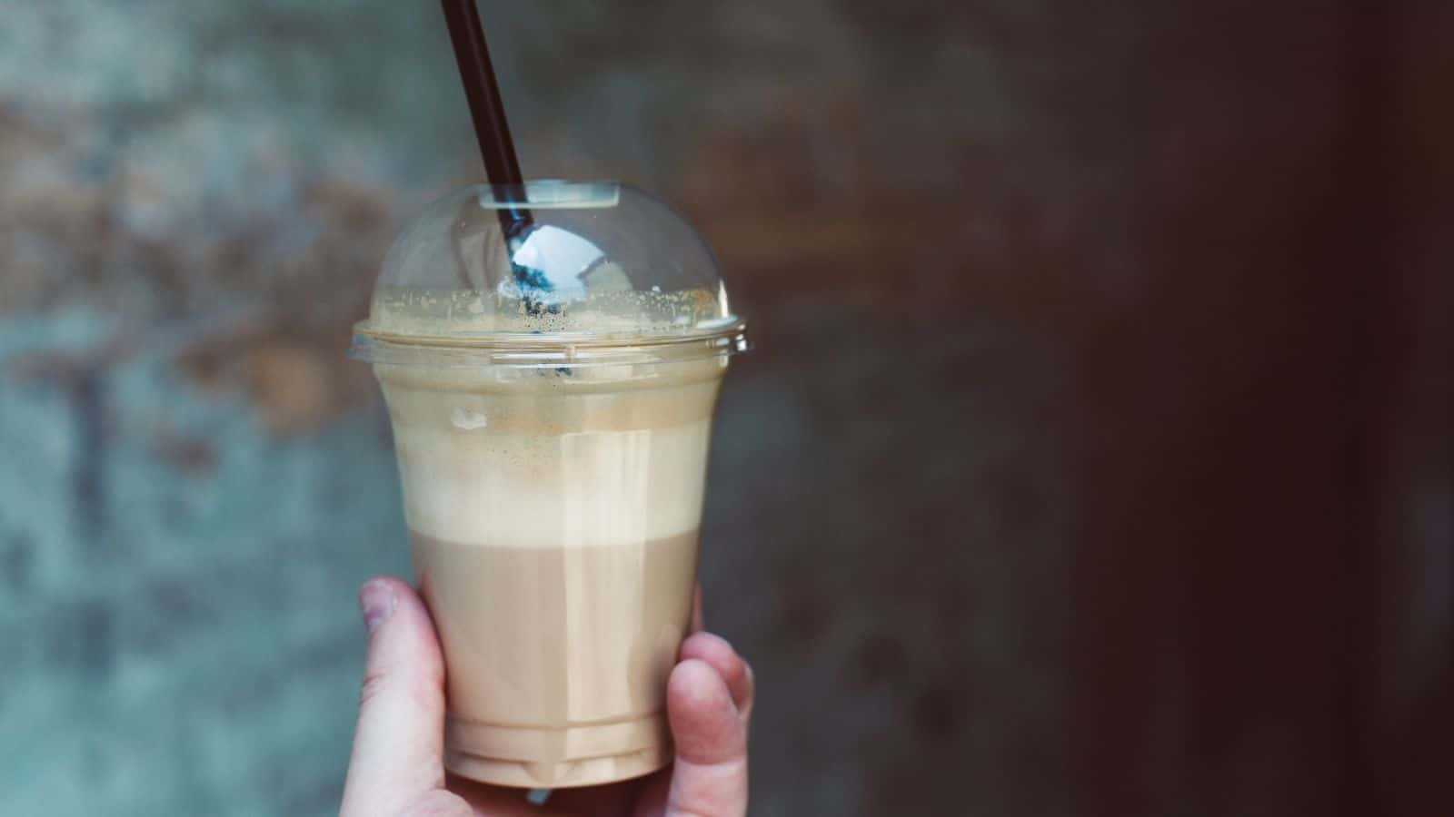 A hand holds a plastic cup with a dome lid and a black straw, containing a layered iced coffee drink. The background is blurred and features a textured wall.