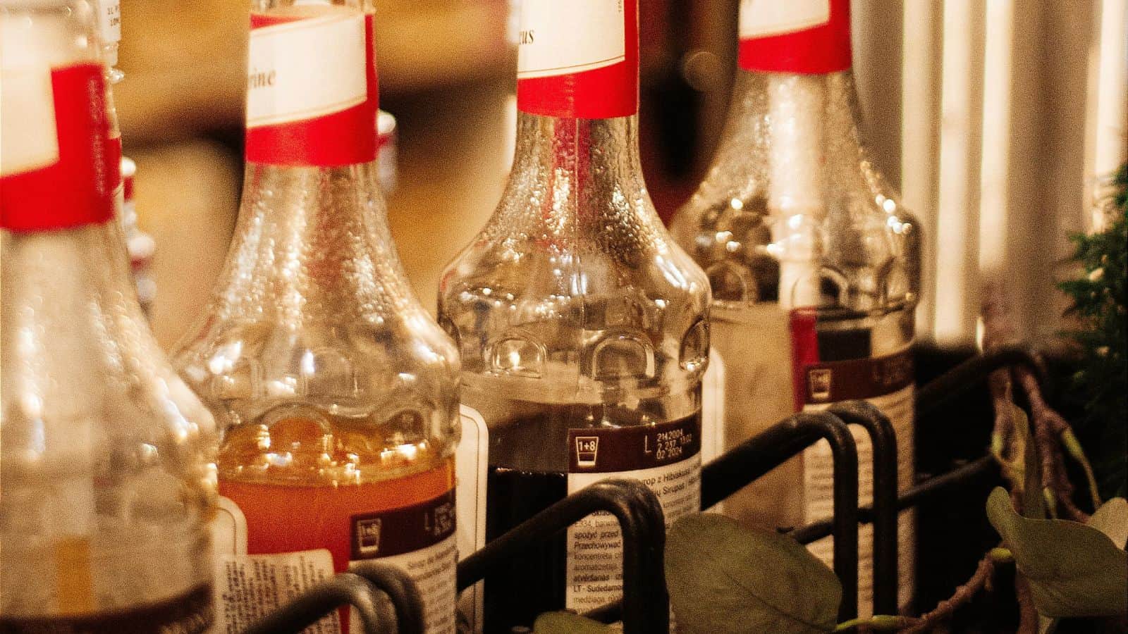 Several glass bottles with red caps and partial labels are lined up in a metal rack. The bottles contain different colored liquids, suggesting various types of beverages. Greenery is visible in the foreground.