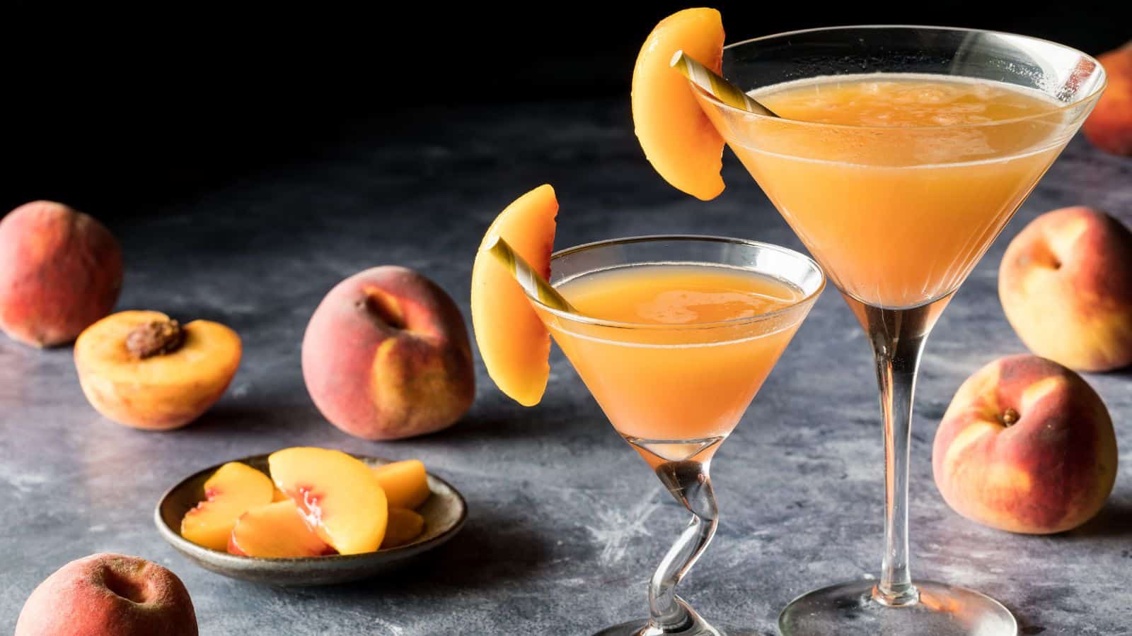 Two martini glasses filled with peach-colored drinks are garnished with peach slices. They sit on a textured surface, surrounded by whole and sliced peaches. The background is dark, highlighting the vibrant colors of the drinks and fruit.