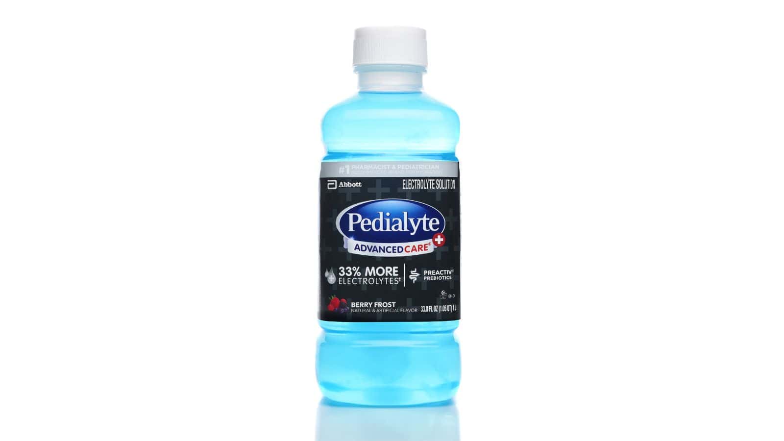 A bottle of Pedialyte AdvancedCare Plus, Berry Frost flavor, with a blue liquid inside. The label highlights "33% more electrolytes" and "preactiv prebiotics." The bottle is standing on a reflective white surface.