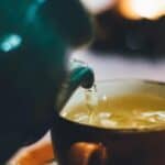 A close-up image of a tea being poured from a green teapot into a ceramic cup. The background is softly blurred and features a warm, glowing light, creating a cozy atmosphere.