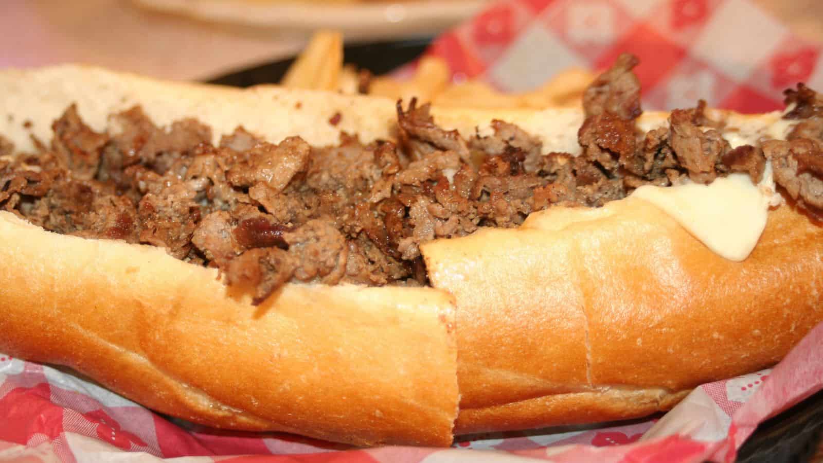 A close-up of a sandwich on a tray, featuring a sliced hoagie roll filled with chopped, cooked meat, likely beef. The sandwich is accompanied by a few visible fries, all on a red and white checkered paper.
