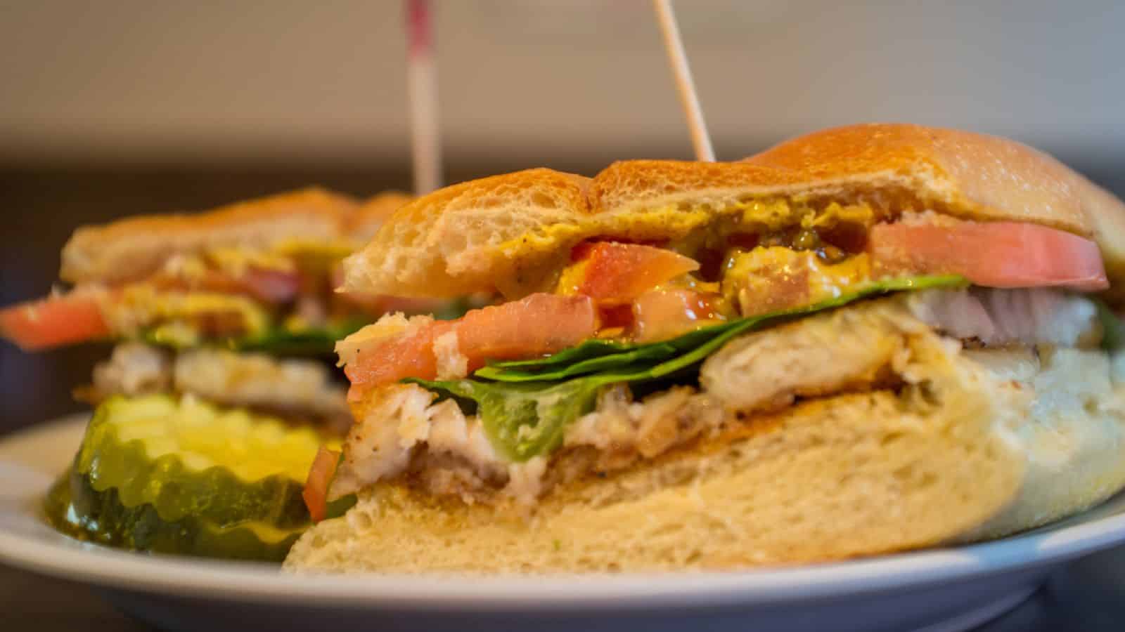 Close-up of a sandwich with sliced chicken, lettuce, tomato, and sauce placed between two halves of a bun, secured with toothpicks. The sandwich is served on a plate with sliced pickles on the side.
