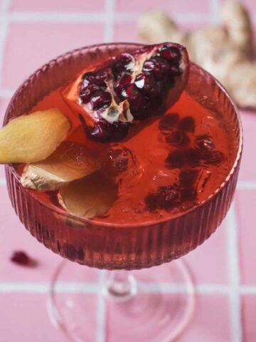 Vibrant pomegranate ginger mocktail in a textured glass, garnished with fresh ginger slices and a pomegranate wedge on a pink tiled background.