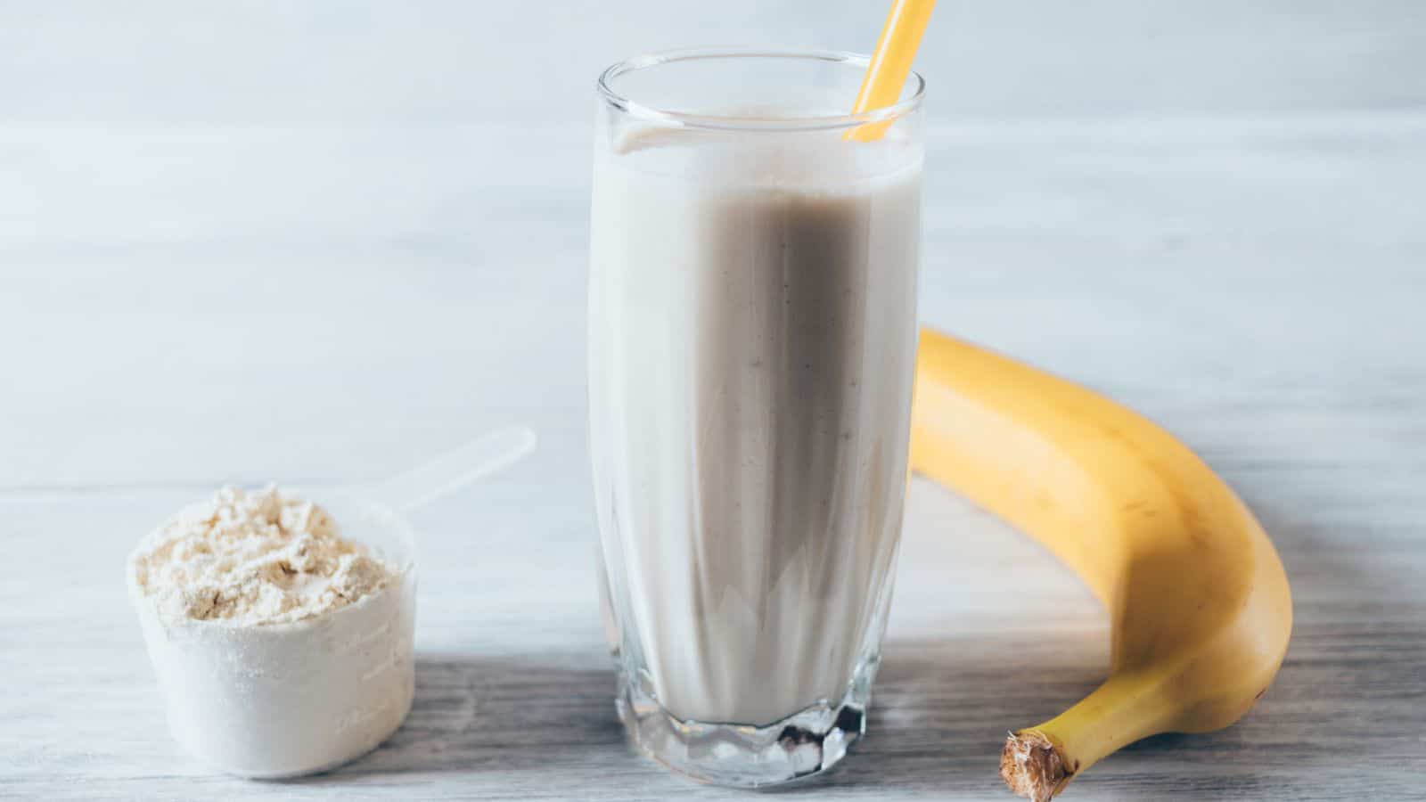 A glass of banana smoothie with a yellow straw is on a table. Next to it is a ripe banana and a scoop of protein powder. The background features a light wooden surface.