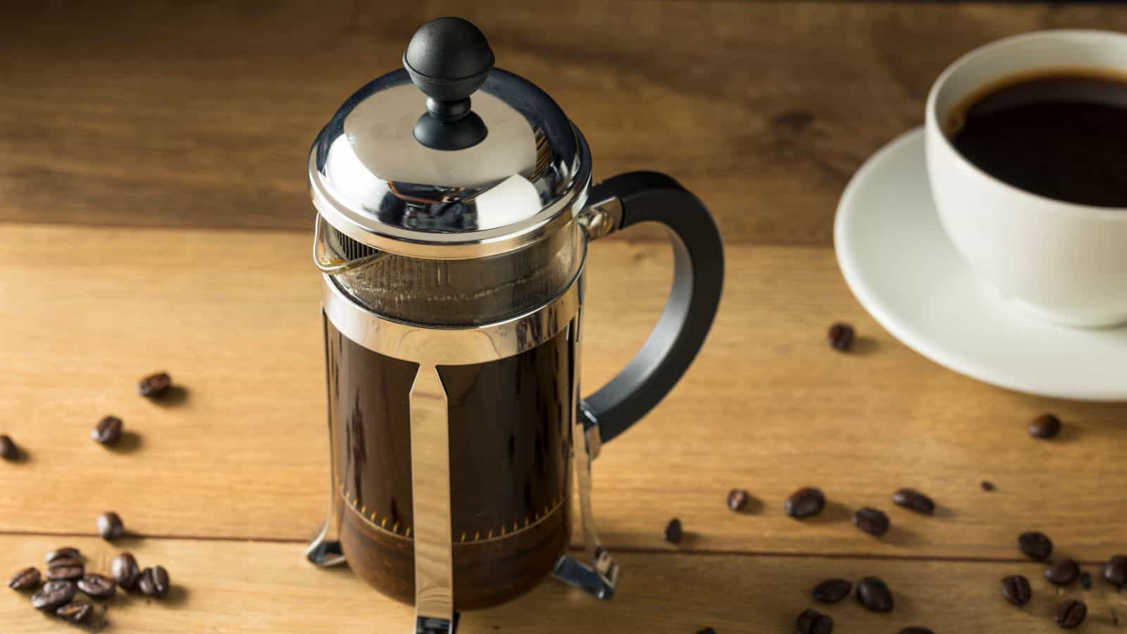 A French press filled with coffee is placed on a wooden table. Coffee beans are scattered around the press, and a cup of black coffee sits on a saucer nearby. The scene conveys a fresh and aromatic coffee preparation setting.