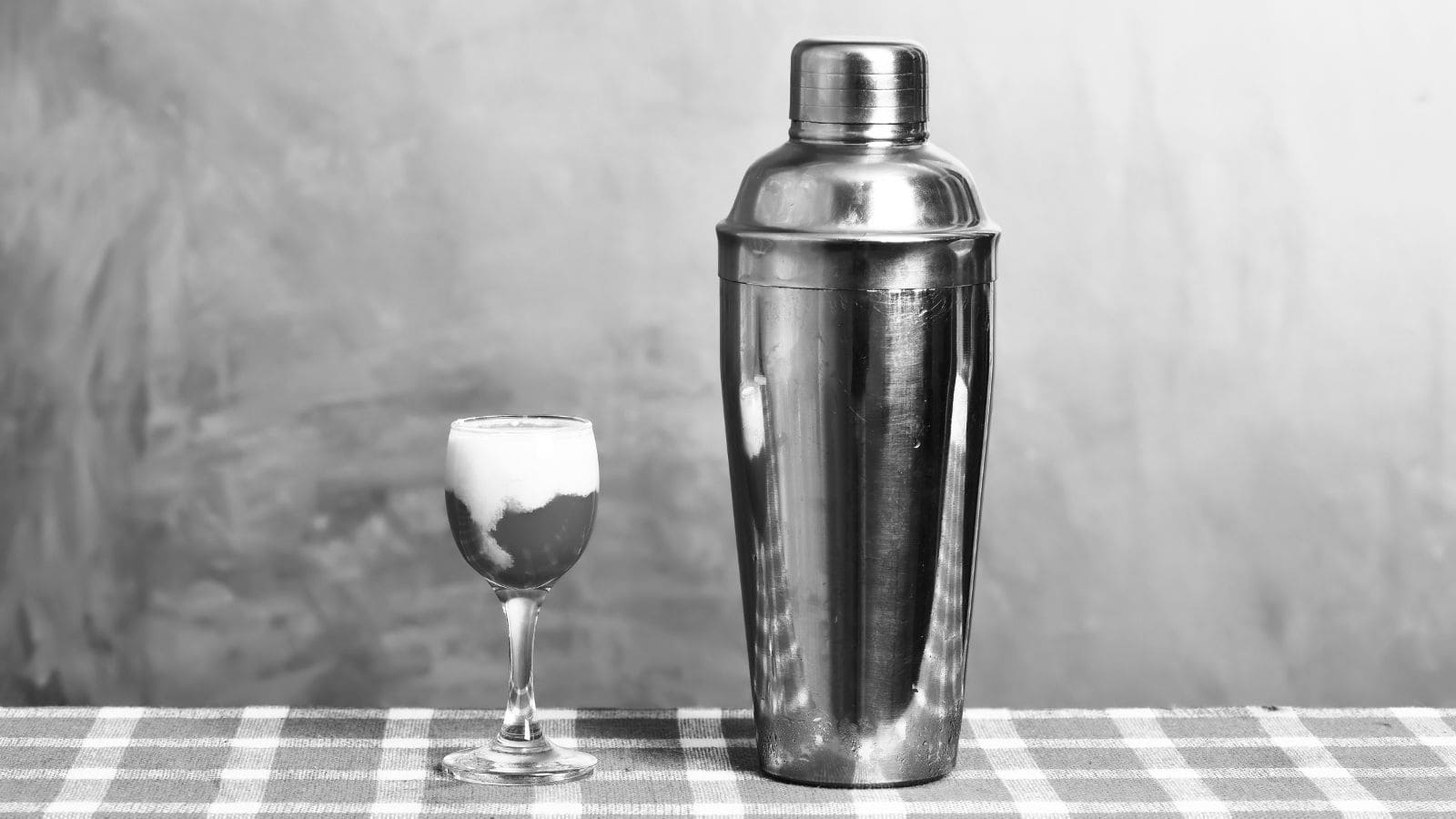 A large metal cocktail shaker is placed on a checkered tablecloth next to a small filled cocktail glass, set against a plain background. The scene is in black and white.