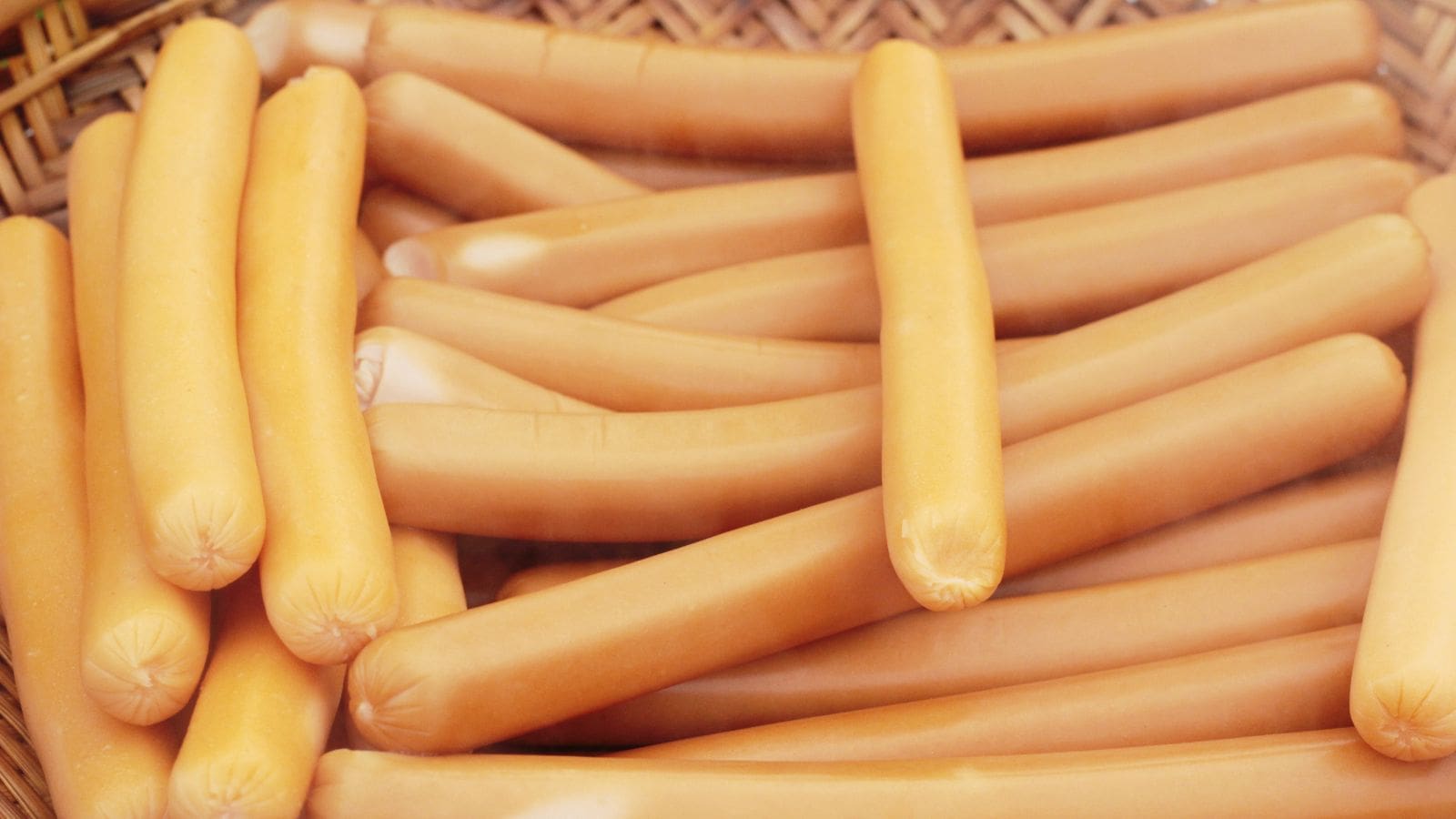 A pile of uncooked sausages is arranged inside a woven basket. The sausages have a smooth, light brown skin and are evenly stacked, covering most of the visible area. The basket background adds a natural texture to the composition.