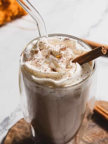 A pumpkin spice cold brew in a glass, topped with whipped cream and a cinnamon stick, with decorative elements in the background.