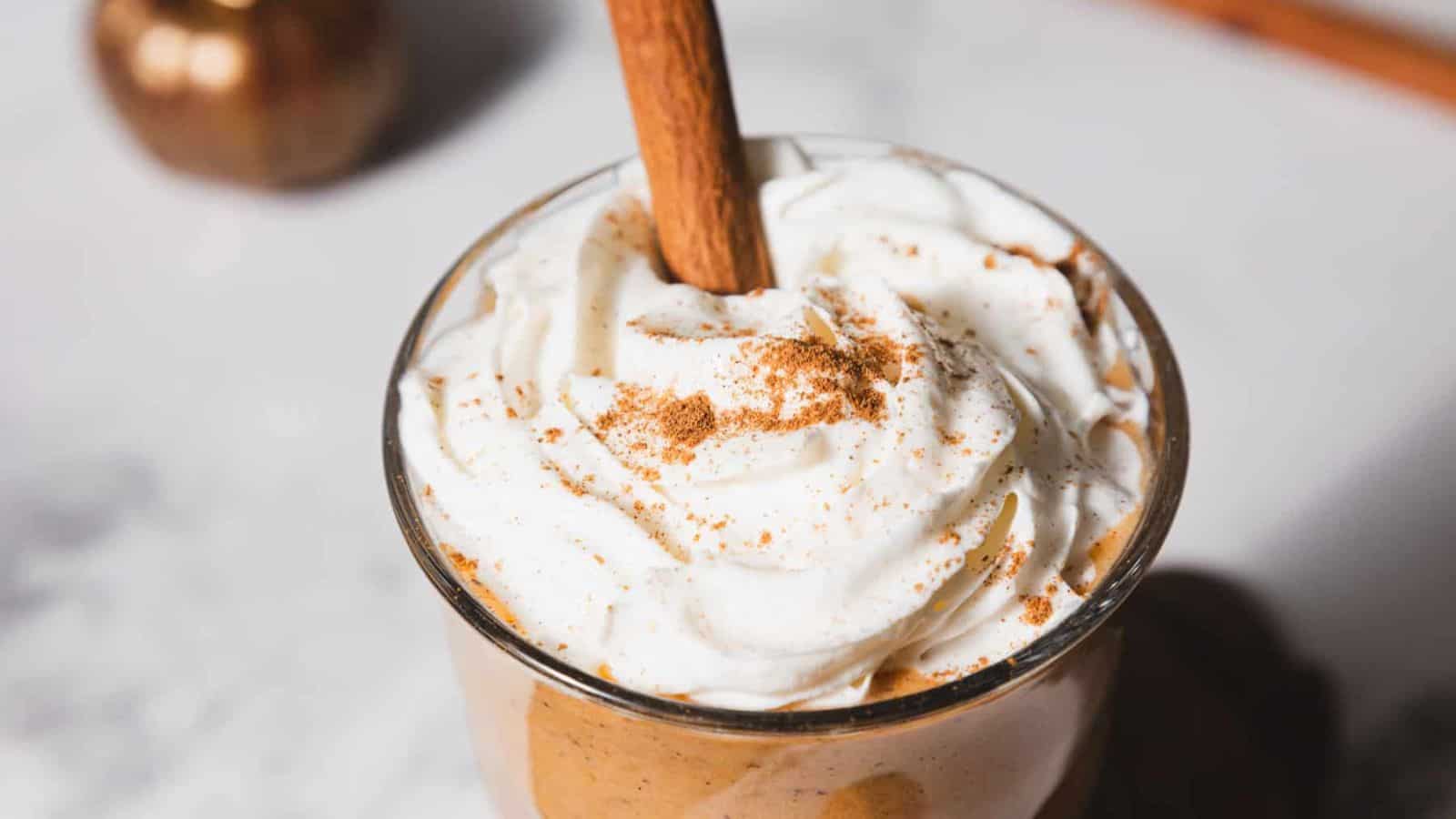 A tall glass filled with a creamy pumpkin spice latte topped with whipped cream and a cinnamon stick. The drink is set on a wooden coaster, and small decorative pumpkins are visible in the background on a marble surface.