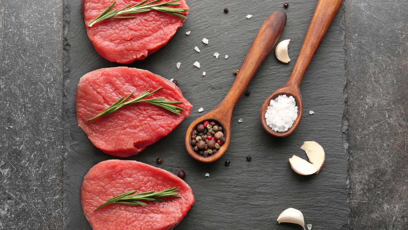 Three raw beef steak pieces on a slate board, each topped with a rosemary sprig. Wooden spoons hold salt and mixed peppercorns. Garlic cloves are scattered around.