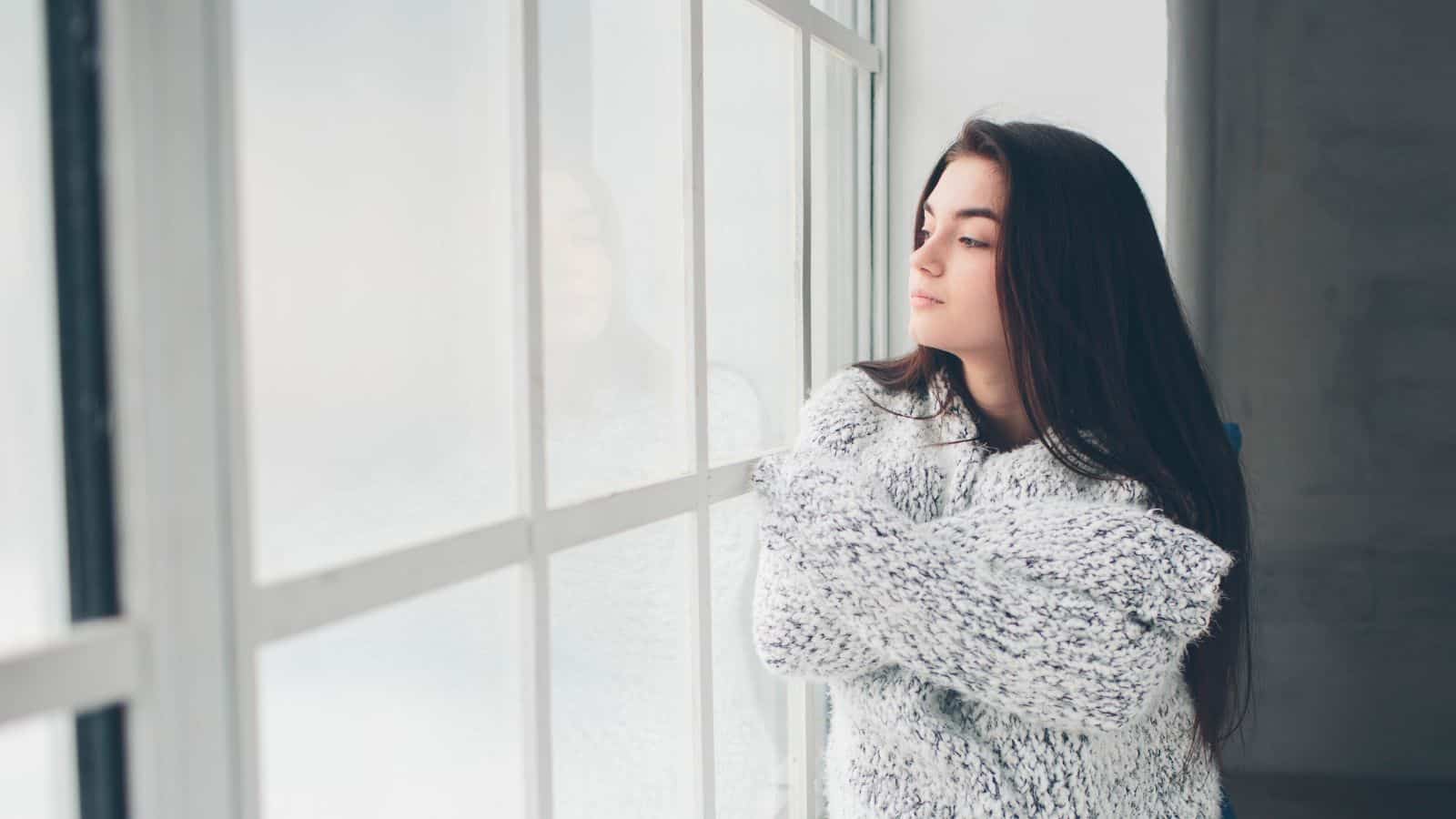 A woman with long dark hair stands indoors, wearing a thick, knitted sweater. She is looking out of a large window, with her arms folded across her chest. The room appears to be bright with natural light streaming in.