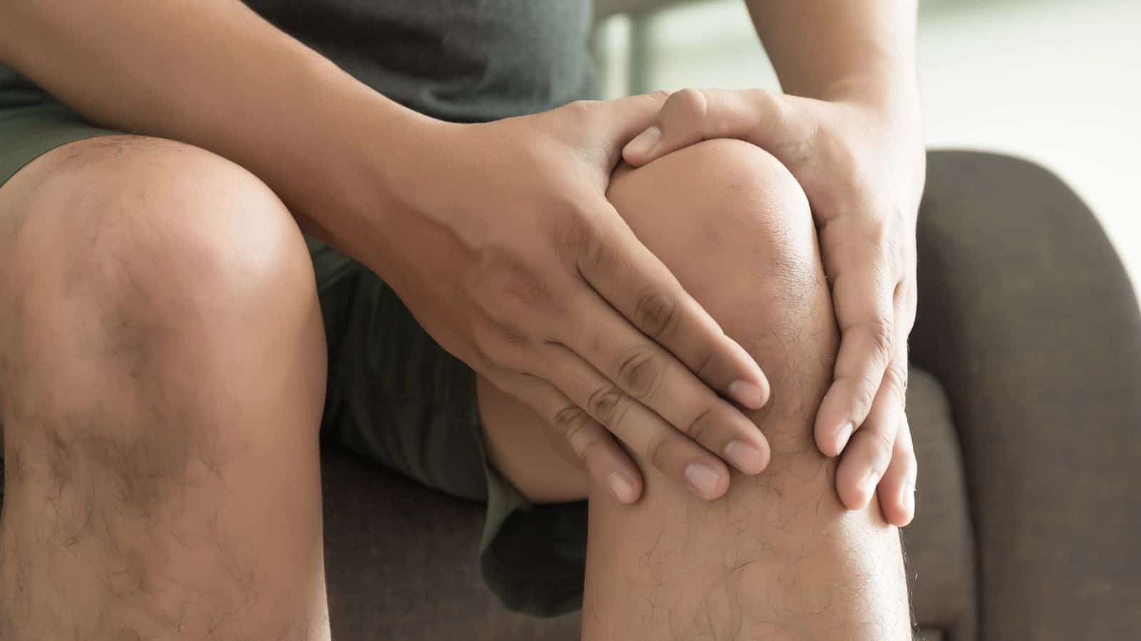 A person sitting on a chair holds their knee with both hands. They are wearing dark shorts and a dark shirt. The background is blurred, focusing attention on the individual's knee.