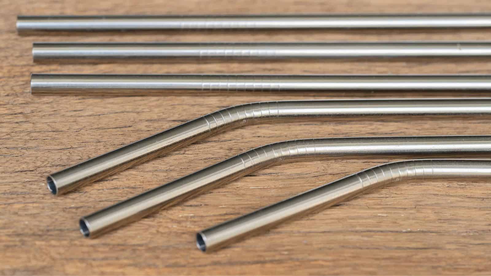 A set of stainless steel straws is displayed on a wooden surface. The straws include both straight and bent designs, arranged neatly in rows.