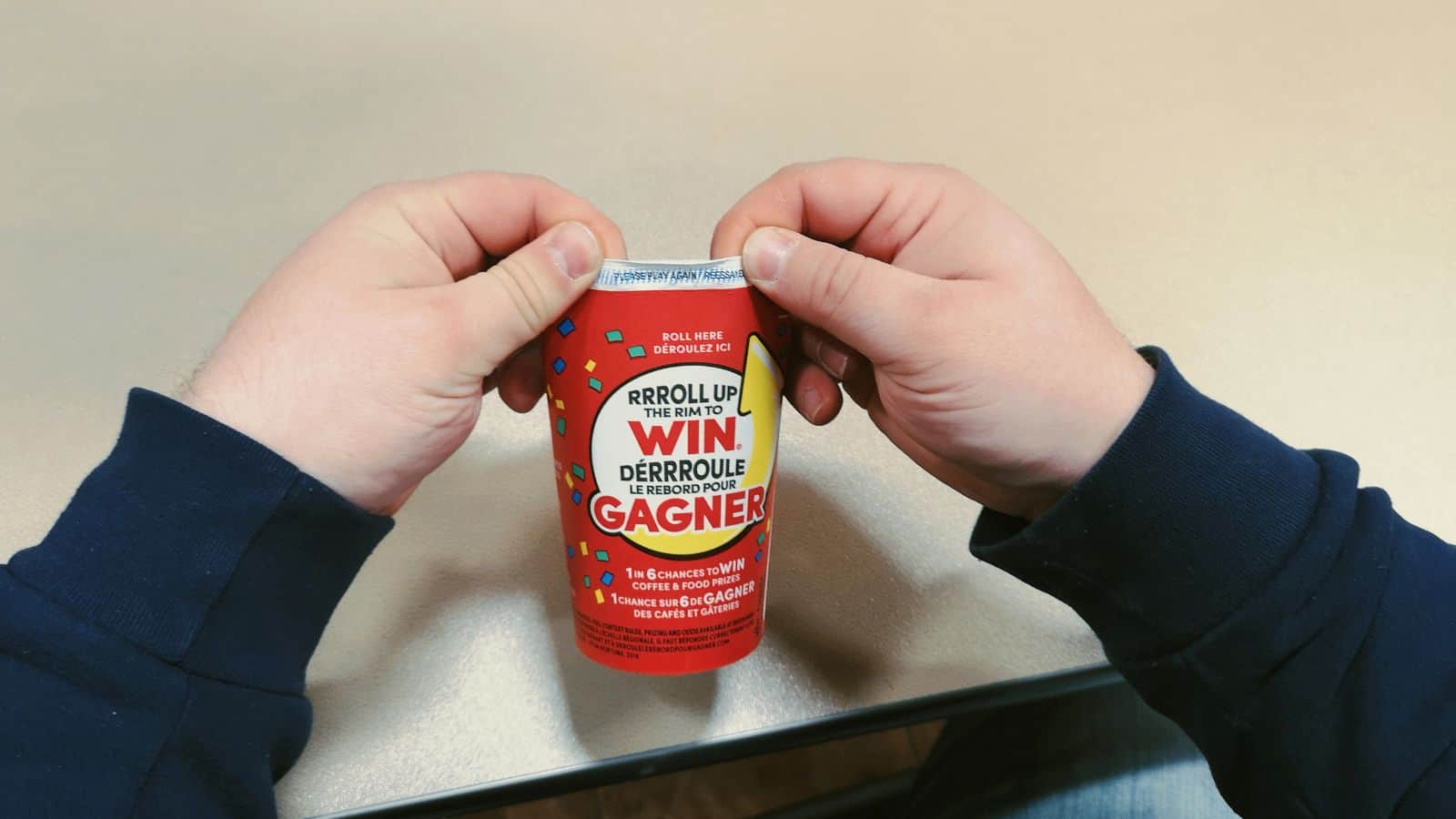 A person wearing a navy blue sweater rolls up the rim of a red coffee cup with a contest promotion saying "Roll up the rim to win" in English and French. The cup is placed on a tan surface.