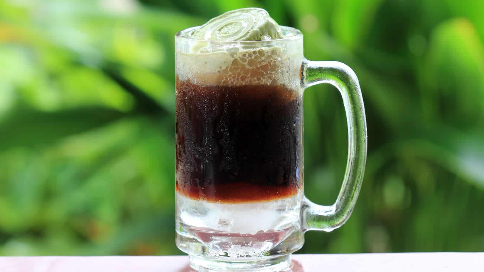 A glass mug filled with dark brown liquid and ice sits on a surface. The beverage has a frothy top, likely from carbonation. The background features green, blurred foliage, suggesting an outdoor setting.