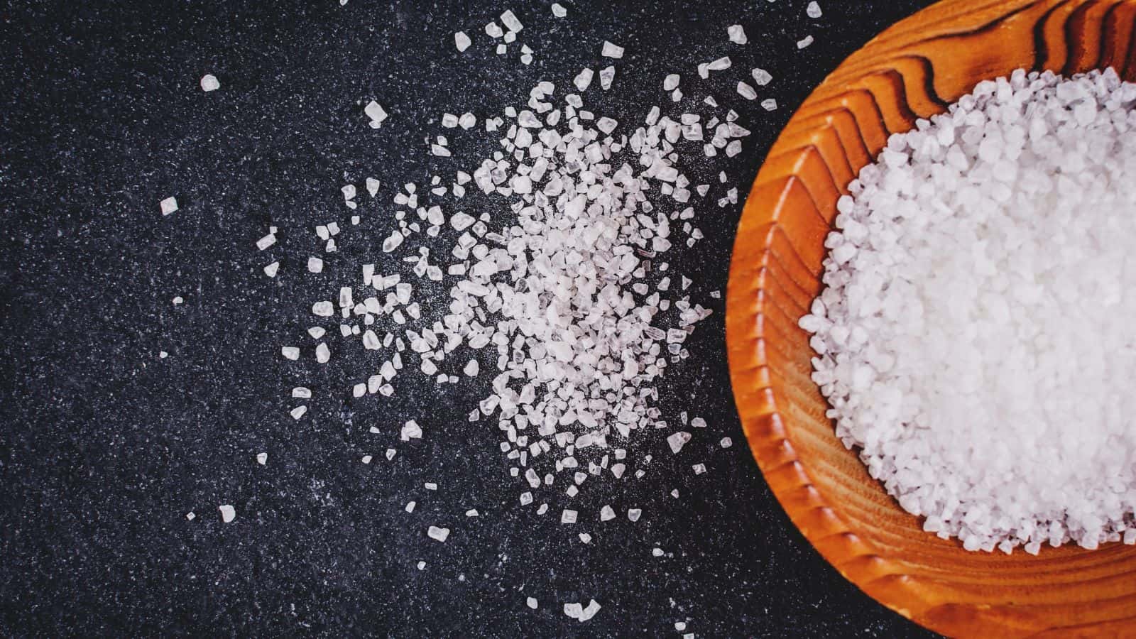 A wooden bowl filled with coarse sea salt is placed on a dark granite surface. Some salt has spilled outside the bowl, scattering across the countertop. The texture of the salt crystals and the wood grain is visible.