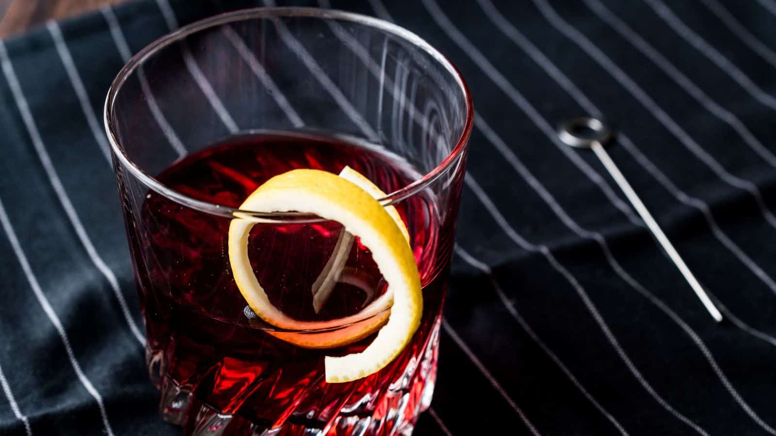 A glass of dark red liquid with a lemon twist garnish is set on a black fabric with white stripes. A metal stick with a circular handle lies nearby.