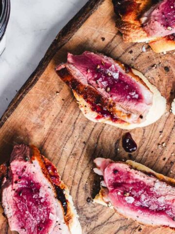 Slices of rare-cooked meat on small pieces of bread are topped with a sprinkling of salt and served on a wooden board. Near the board is a jar filled with dark liquid, possibly jam or sauce. The setting is a marble countertop.