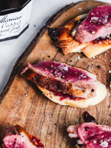Slices of beef on toasted bread with a dark berry sauce are arranged on a wooden serving board. A jar of Bonne Maman blackcurrant jelly is nearby. Another piece of plain toasted bread is on a plate to the side.