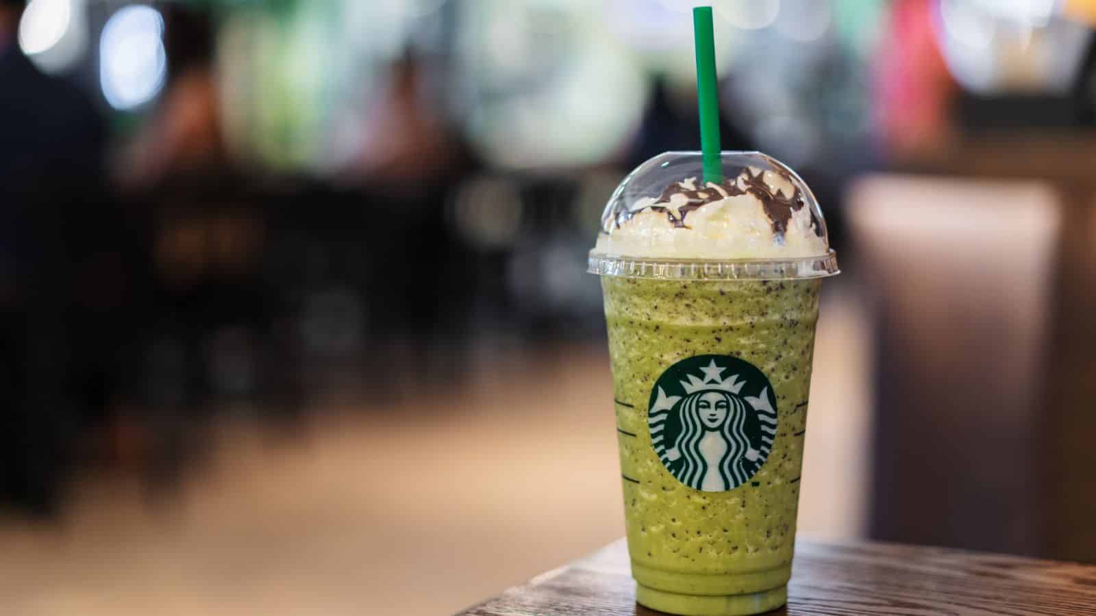 A green Starbucks Frappuccino topped with whipped cream and chocolate drizzle is placed on a wooden table. The background is a blurred image of a cafe interior.