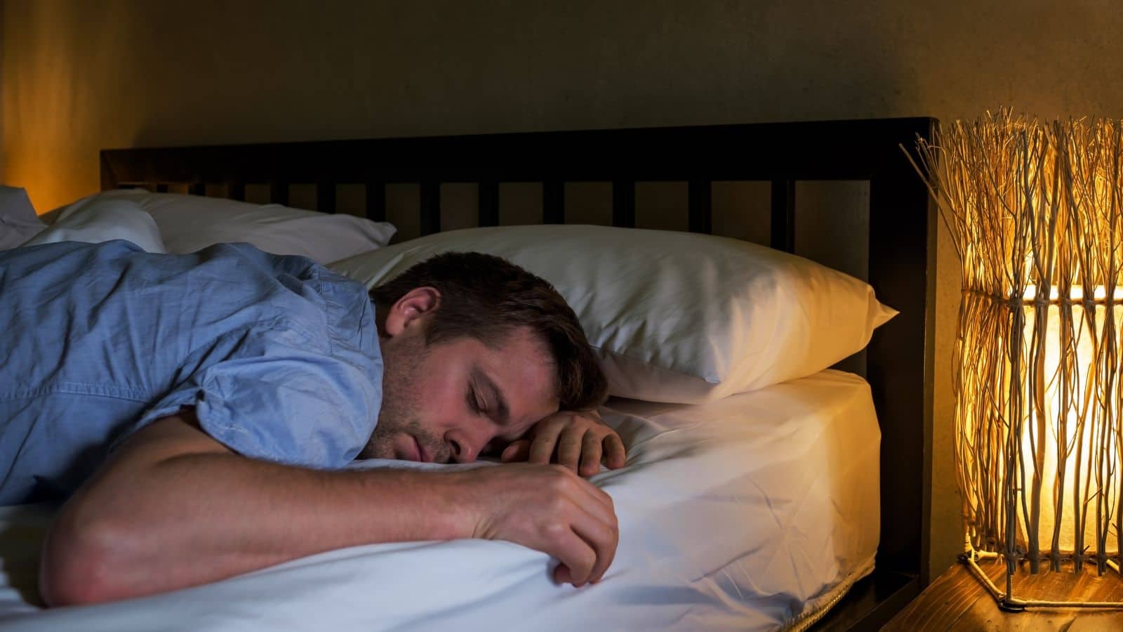 A man is lying face down on a bed, sleeping. He is wearing a blue shirt. The bed has white sheets and pillows. A decorative lamp with a wicker shade emits a warm glow on the right side of the image.