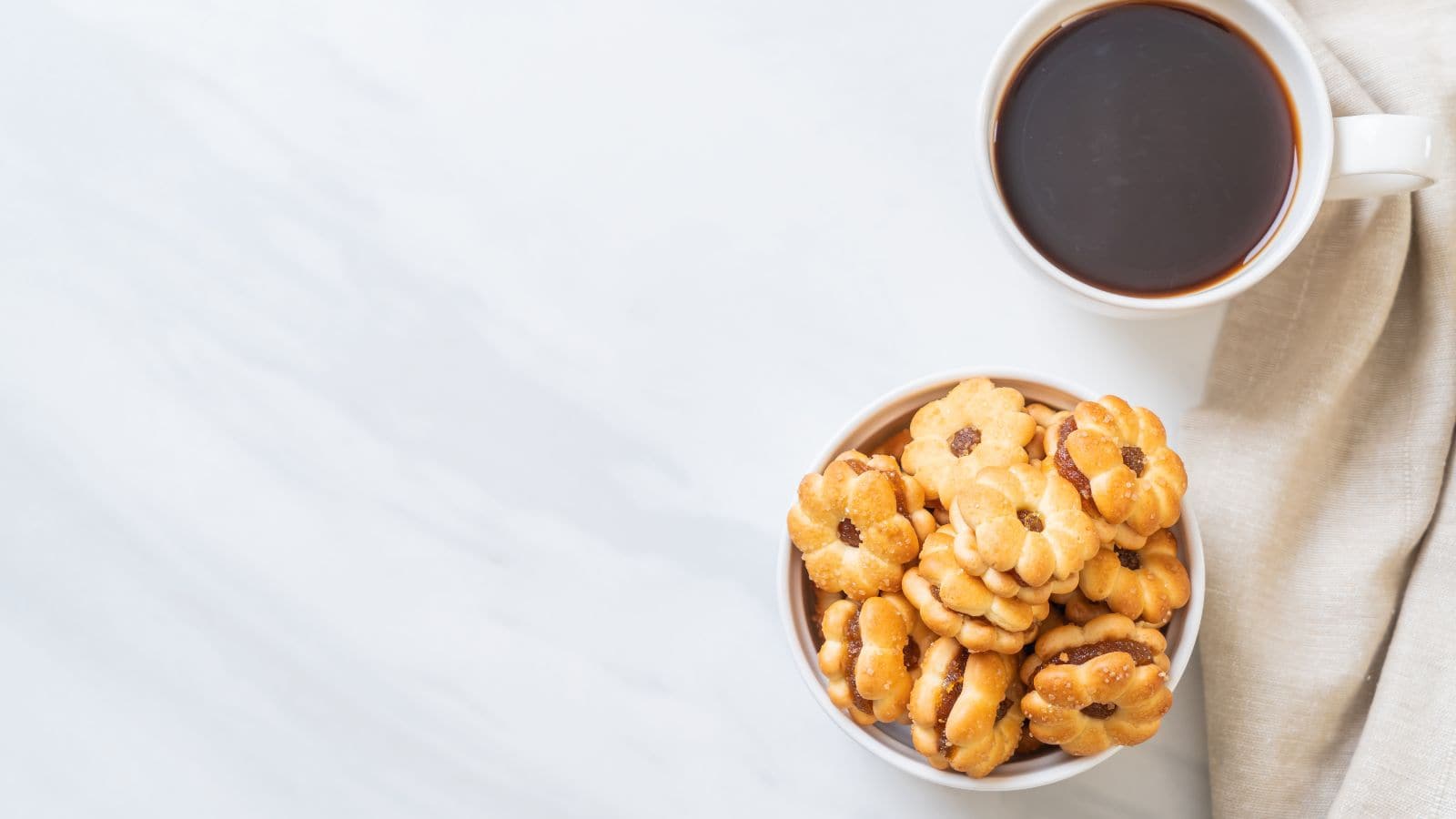 A white cup filled with black coffee sits on a light surface next to a bowl of flower-shaped cookies with a small filling in the center. A beige cloth partially drapes beside the cup. The setting is simple and clean.