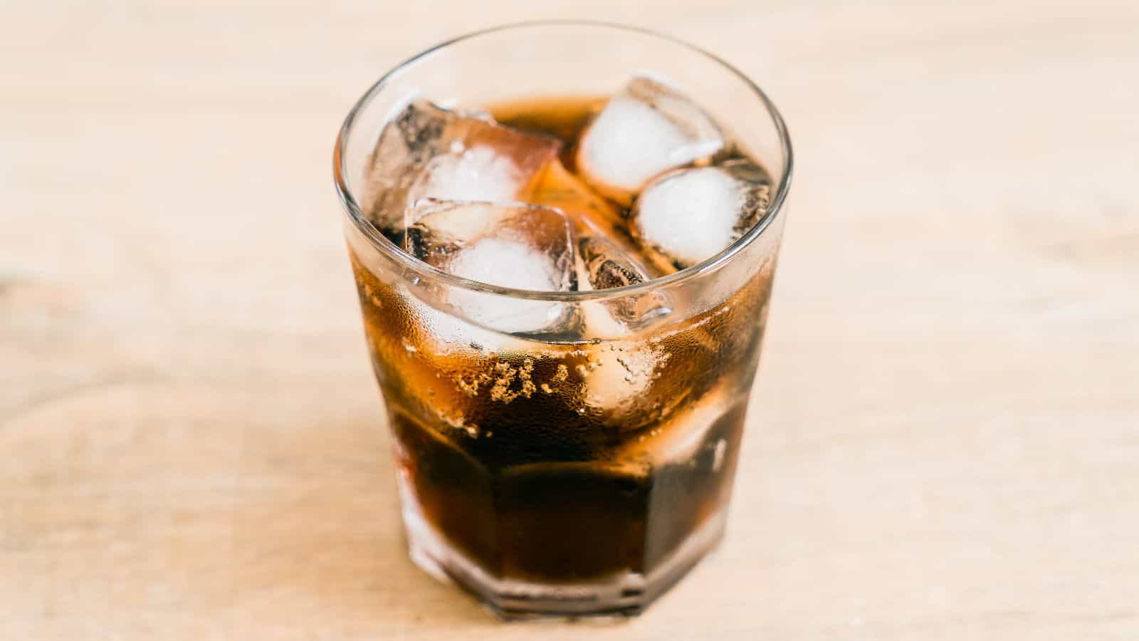 A glass filled with a dark carbonated beverage and ice cubes sits on a light wooden surface.