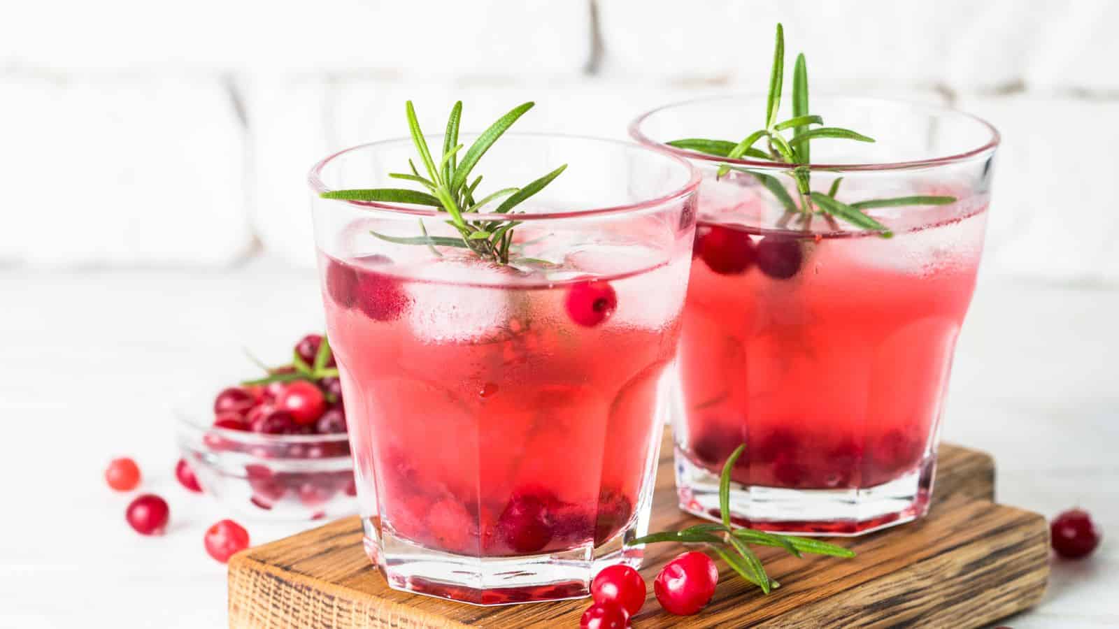 Two glasses of cranberry cocktails on a wooden board, garnished with sprigs of rosemary. Ice cubes and cranberries float in the pink-hued drinks. A small bowl with cranberries is in the background.